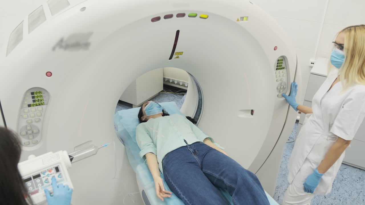 la paciente está siendo sometida a una tomografía computarizada o resonancia magnética bajo la supervisión de dos radiólogos calificados con máscara y guantes en una clínica médica moderna. la paciente está acostada en una mesa de tomografías computarizadas o mri