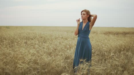 Handsome-young-woman-in-a-long-blue-dress-standing-in-golden-wheat-field-trying-the-wheat's-stem,-touching-her-hair-and-enjoying