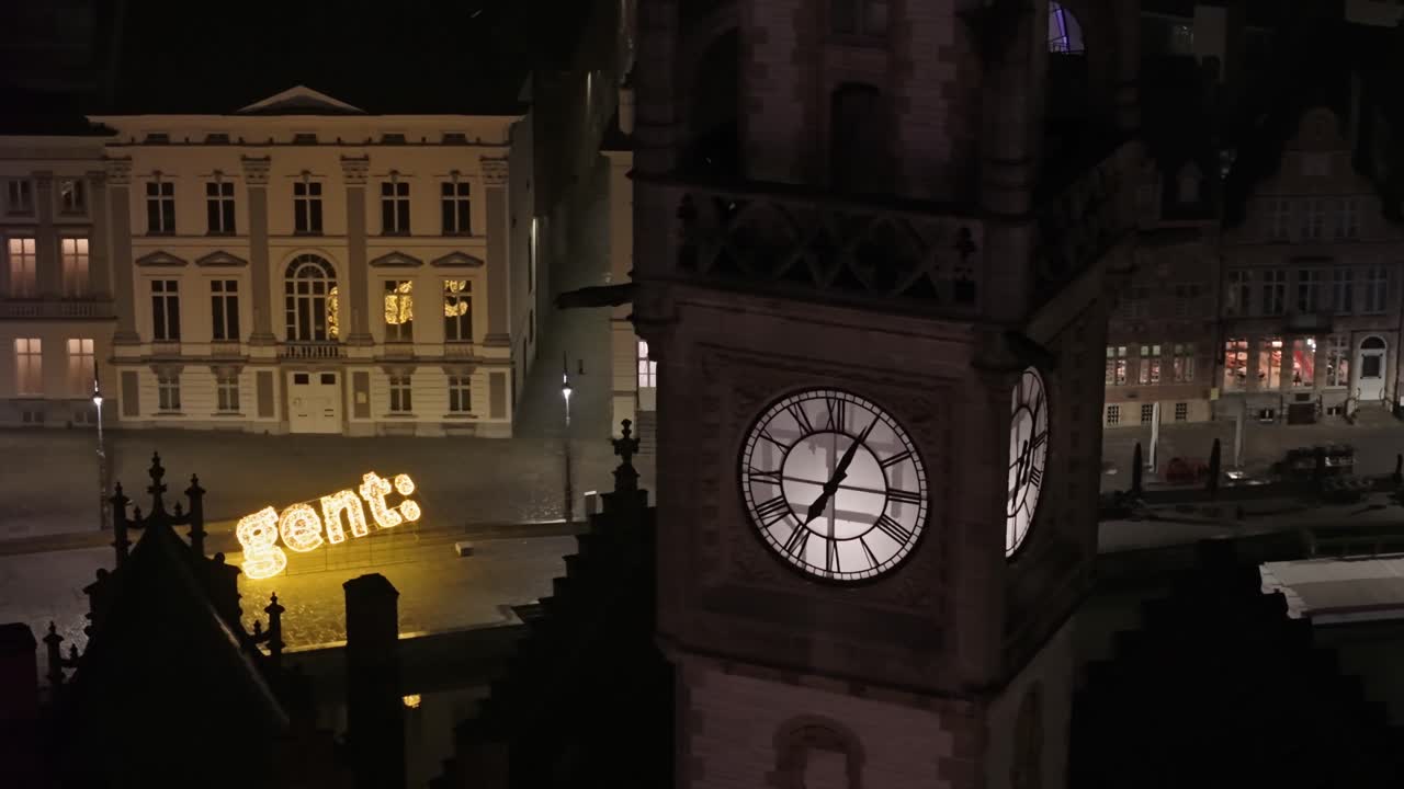 Drone footage of the illuminated clock tower in Ghent, revealing the canal, waterside buildings, and the city sign in an atmospheric urban scene