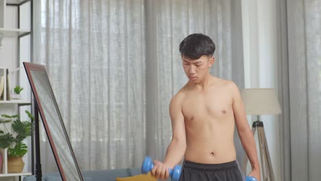 아시아 십대 소년 은 집 에서 셔츠 를 벗고 운동 하는 동안 덤벨 으로 훈련 하고 거울 을 보고 있다