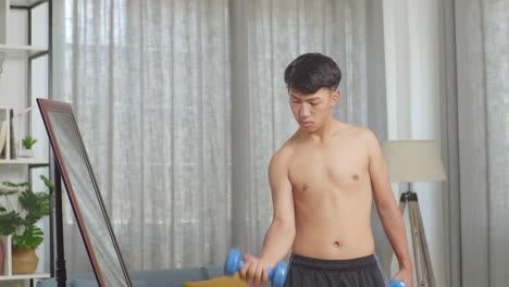 asian teenager boy training with dumbbells and looking at the mirror while doing shirtless workout at home