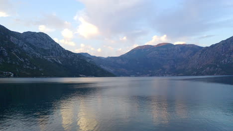 the bay of kotor in montenegro, with its still waters, surrounded by high mountain range, reflected in the water, white clouds above the horizon, late afternoon sunlight shining over the peaks