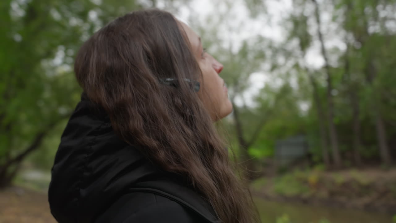 Donna bianca che fotografa con il telefono gli alberi sulla riva del fiume, di spalle rispetto alla camera vicino alla riva cosparsa di foglie, cappotto e cappuccio neri, attenta osservatrice che inquadra il ponte e il riflesso sull'acqua, atmosfera curiosa