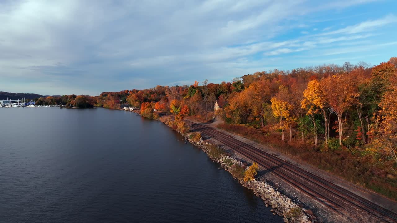 An aerial view over the Hudson River by empty train tracks on a sunny day in autumn. The camera dolly in along the tracks and colorful trees.