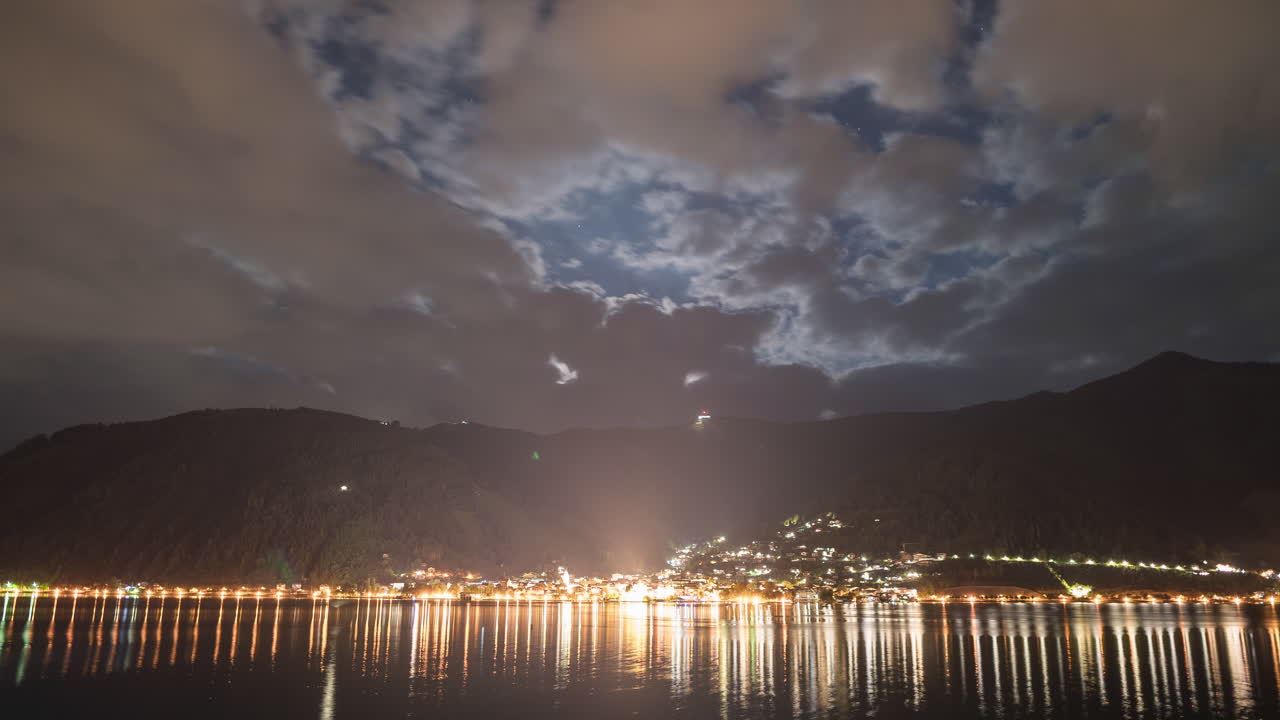 Night sky and city timelapse across the lake in Zell am See, Austria. Stars and moon crossing the sky in between cloud movement