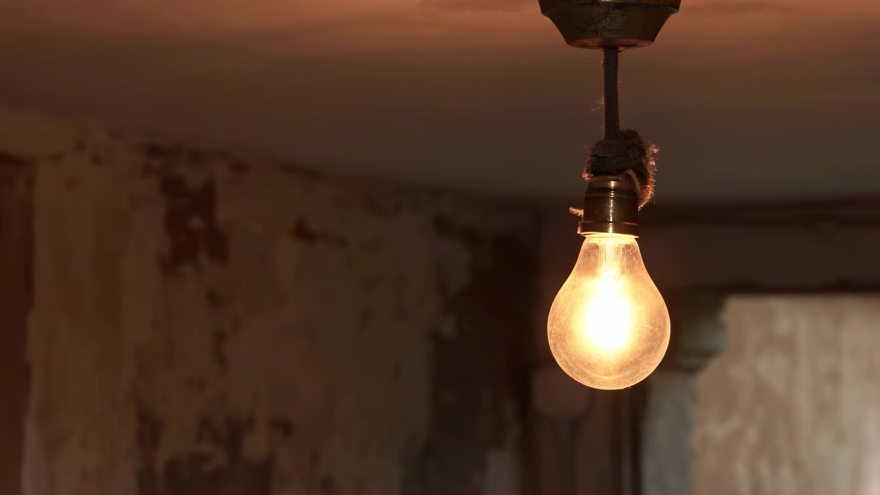 Vista ravvicinata di una singola lampadina a incandescenza appesa che cambia tra bagliore luminoso e stato spento all'interno di una stanza rustica e usurata, con intonaco scrostato, pareti macchiate, portalampada ossidato e cavo rivestito in tessuto