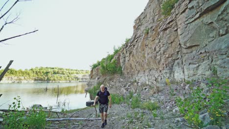 adult tourist is walking along the bank of the lake with a stick in his hand and with a backpack on his shoulders on the background of high rocks in the summer