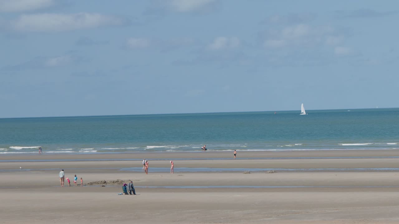 Wide shot of people strolling on sandy shore, sailboats in distance, bright daylight, tranquil mood