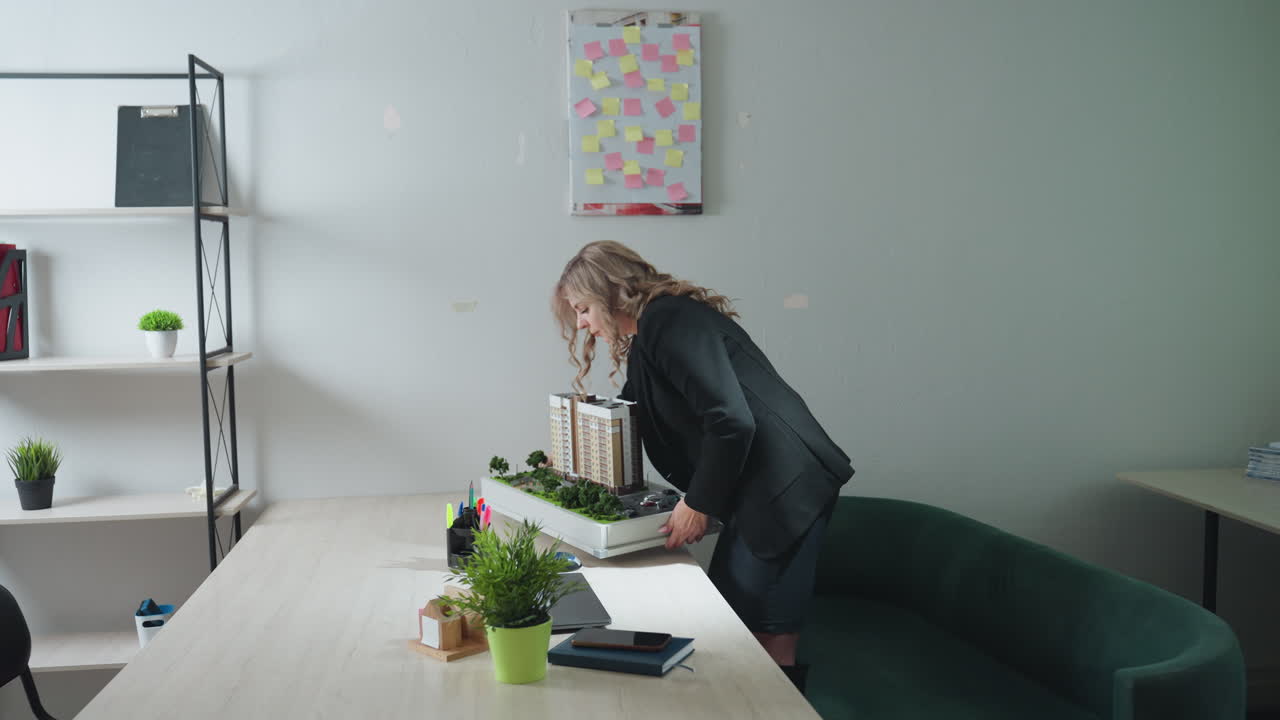 Real estate agent carrying architectural building model walks toward desk and drops it in modern office workspace with minimalist decor, featuring desk plant, bookshelves and green couch