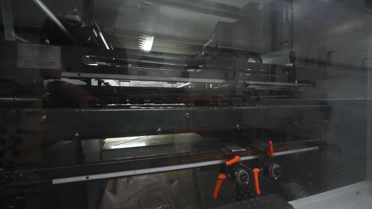 Frontal slightly angled view of industrial machinery behind protective glass, with dense metal parts, reflective surfaces, overhead glare, blue panel accents, and utilitarian factory atmosphere