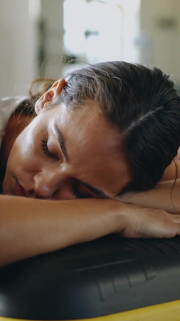 Vertical video: Lying woman in gray top post-workout breathing and shifting on aerobic step in gym