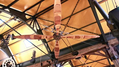 a giant mechanically operated wooden ceiling fan rotates in a huge warehouse with heavy steam punk influence in the design