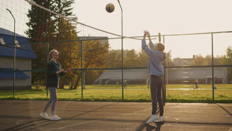 youngsters playing volleyball on court with ball, background featuring open field and blurry view of someone pushing bicycle and other people walking in park