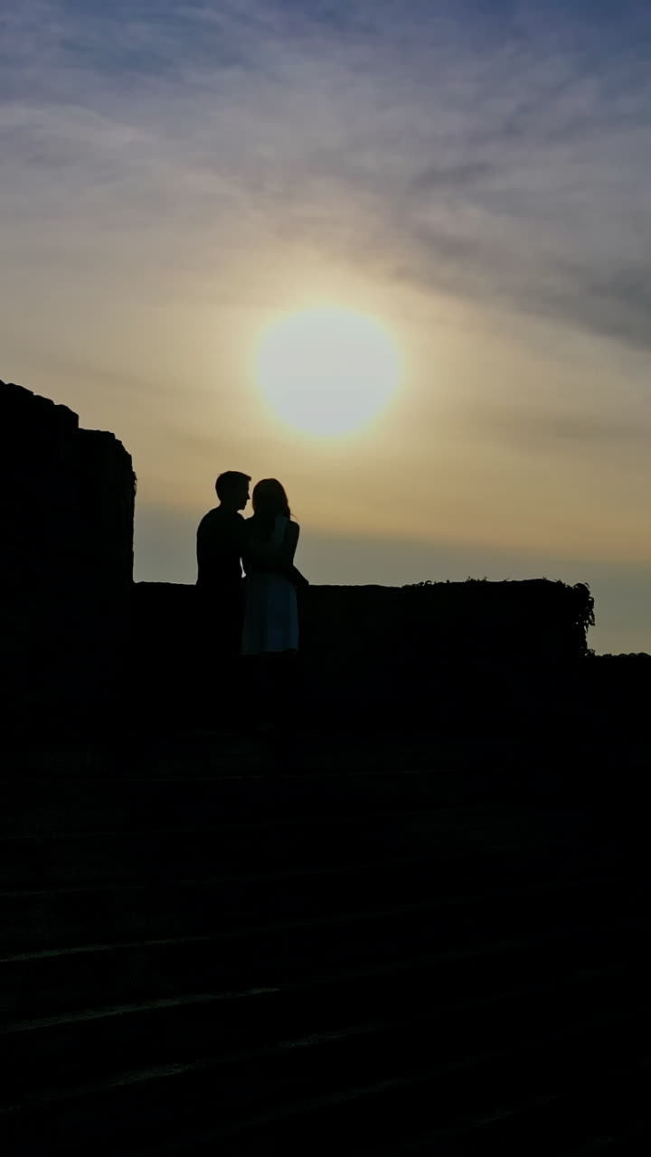 Romantic couple in love spending time together at sunset. Young man kisses girlfriend standing in a castle over the water in the evening. Vertical video