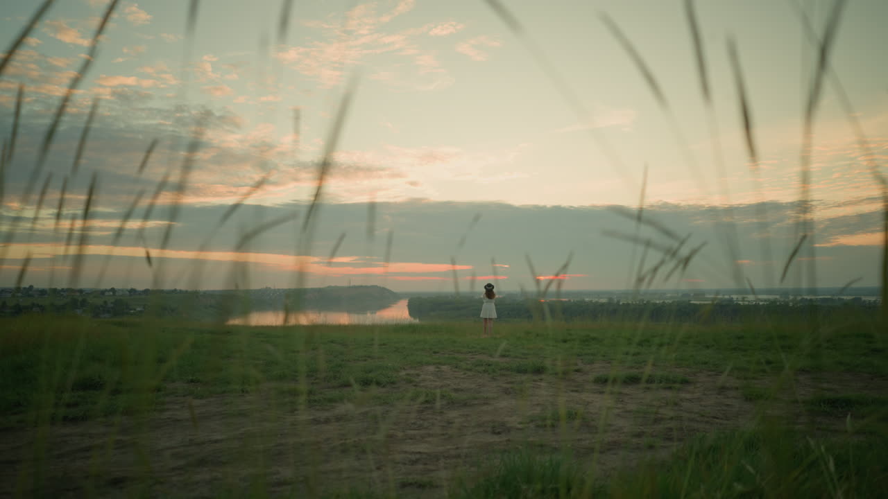 색 드레스와 모자를 입은 여자가 먼 곳에서, 해가 지는 동안 조용한 호수 의 잔디에서 사려 깊게 서있는 모습. 그녀는 평온하고 반영적인 순간을 포옹하여 지평선으로 쳐다보고 있습니다.