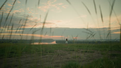 a distant view of a woman in a white dress and hat, standing thoughtfully in a grassy field by a tranquil lake at sunset. she gazes into the horizon, embracing the serene and reflective moment