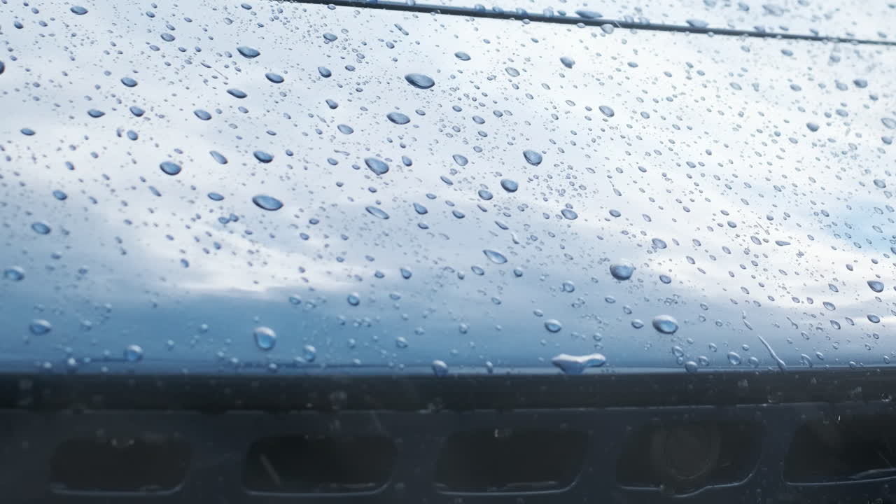 Close up of blue sports car hood with raindrops and reflections of sky and clouds. Cinematic rainy detail of luxury vehicle front and grille