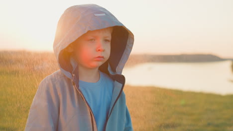 un niño mira pensativo al atardecer mientras está de pie en un campo cerca de un río.