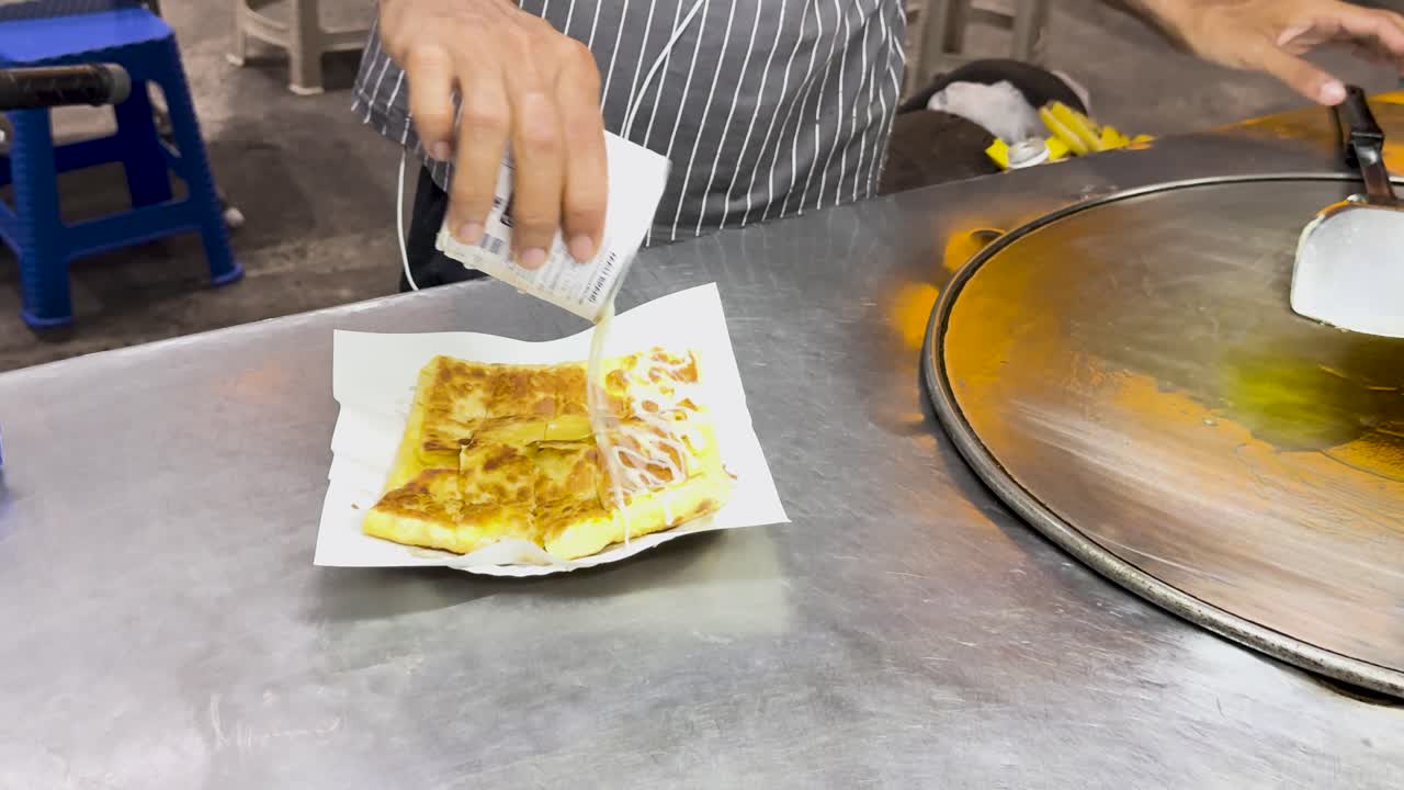 A street vendor in Phuket prepares Thai roti, drizzling condensed milk over a freshly cooked flatbread on a hot griddle