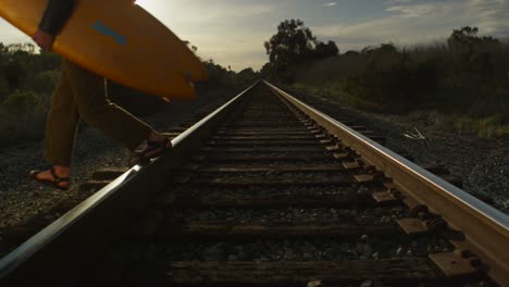 Ein-Surfer-Trägt-Sein-Board-über-Bahngleise,-Während-Er-Zu-Einem-Abgelegenen-Surfspot-In-Einer-Küstenregion-Wandert