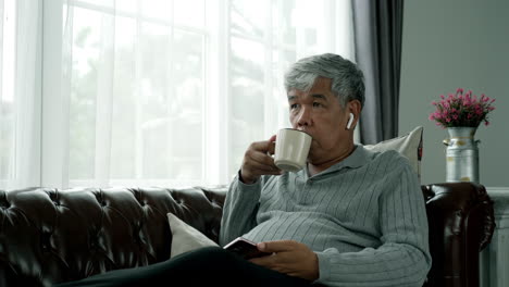 active senior man relaxing at home working, wearing bluetooth earbud enjoy listening to music from a mobile phone sitting comfortably on a sofa