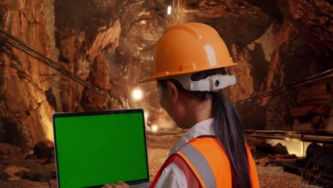 close up back view of asian female engineer with safety helmet working on a green screen laptop and looking around in underground mine tunnel