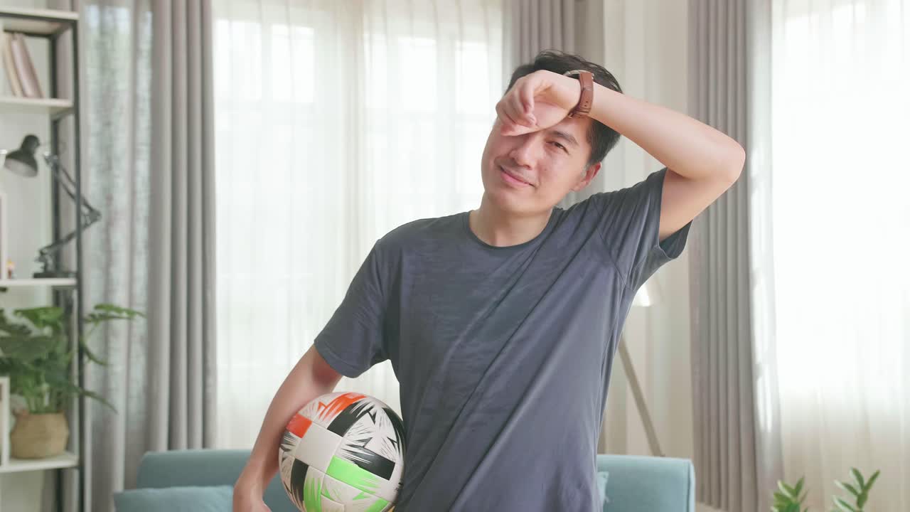 Asian Man Holding Ball Under His Arm And Wiping Sweat From Forehead In Living Room