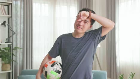 asian man holding ball under his arm and wiping sweat from forehead in living room