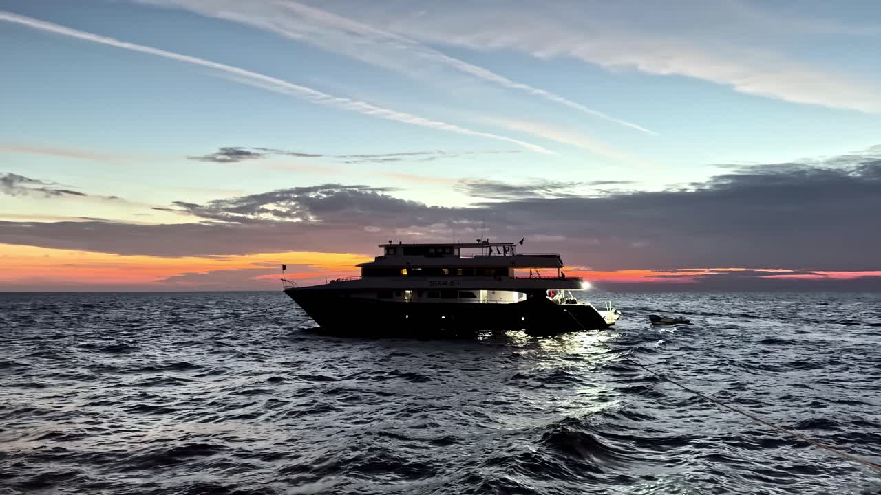 Grande yacht di lusso nero con tender che naviga onde oceaniche calme durante il tramonto nell'ora d'oro, con cielo arancione vibrante e nuvole blu drammatiche, catturato da una prospettiva a basso livello dell'acqua.