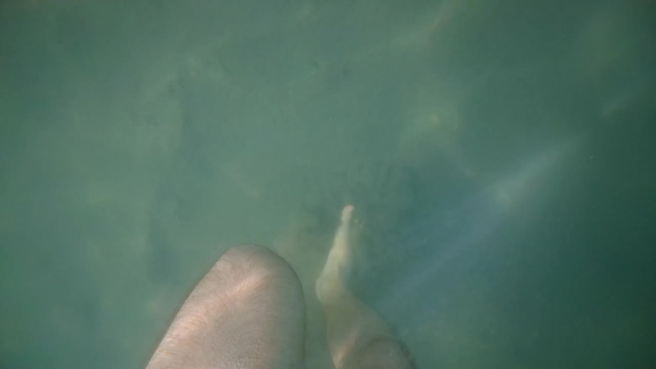 Underwater close up of a man's foot standing in clear shallow sea water, with soft sunlight reflections moving across the sandy seabed