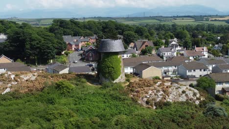 Melin-Wynt-Y-Craig-disused-Llangefni-windmill-ivy-covered-hillside-landmark-aerial-descending-view-to-reveal-Welsh-Snowdonia-mountains,-Anglesey