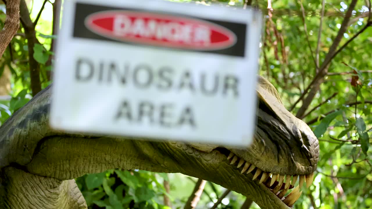 Animatronic raptor head moves through lush greenery as a warning sign swings into view. Daylight, handheld camera, playful and suspenseful mood