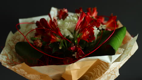 Girando-Alrededor-De-Un-Gran-Ramo-De-Flores-Rojas-Y-Blancas-Y-Elementos-Decorativos-Verdes-En-Un-Jarrón-De-Vidrio-Decorado-Con-Papel-Sobre-Un-Fondo-Oscuro