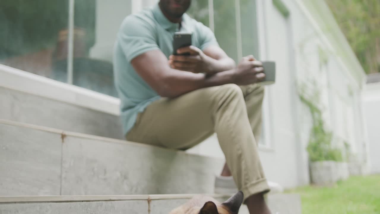 Happy african american man sitting with smartphone and drinking coffee in garden