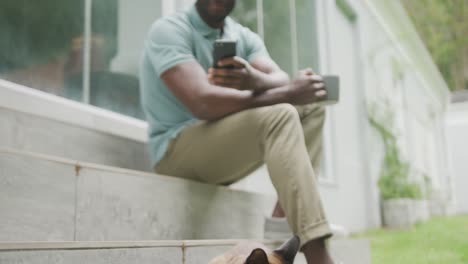 happy african american man sitting with smartphone and drinking coffee in garden