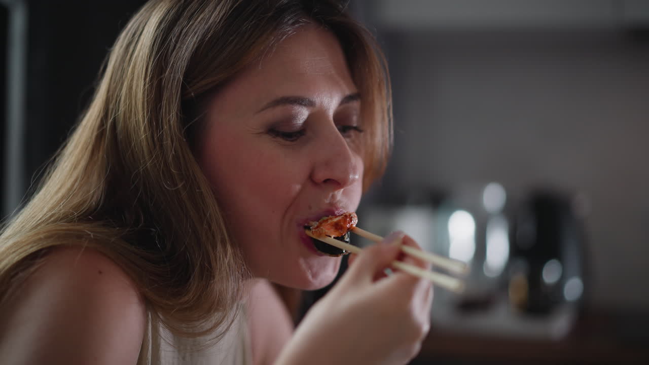 Close up of elegant young woman using chopsticks to eat seasoned meat sushi roll indoors, eyes partially closed while savoring taste, soft lighting and blurred background in kitchen