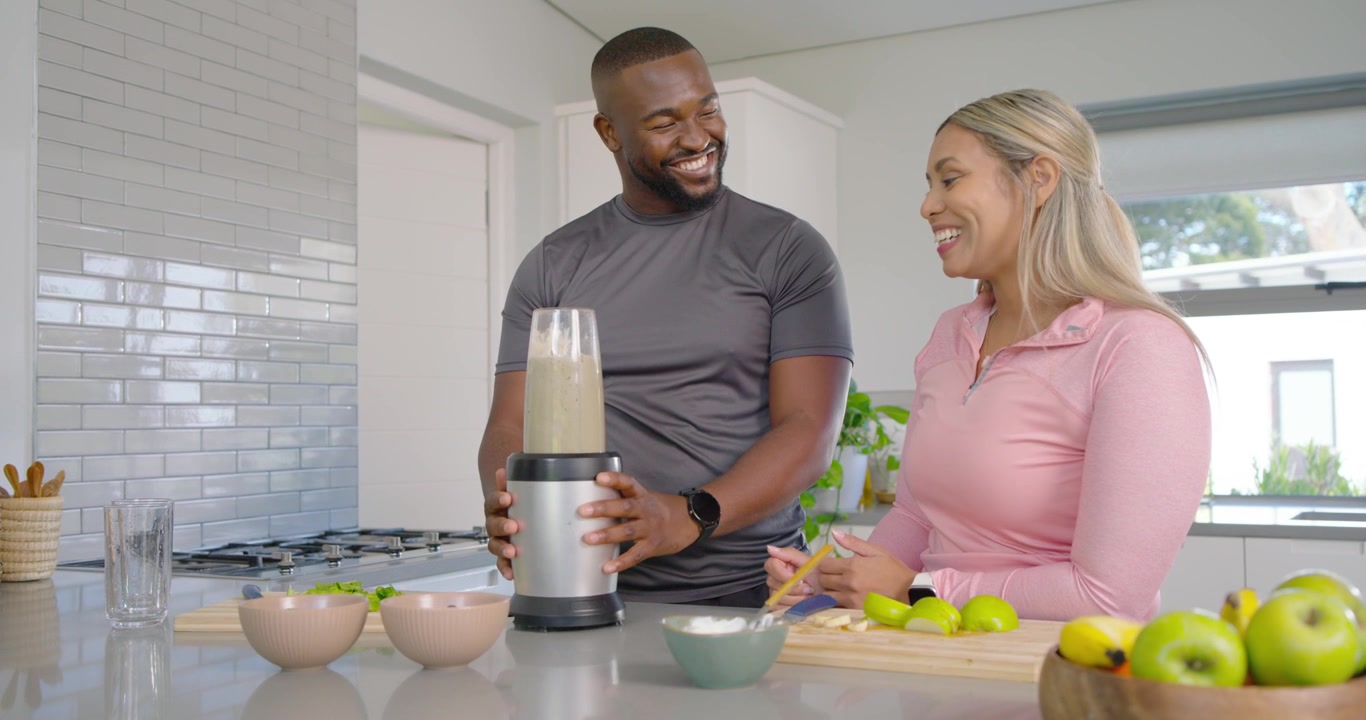 Couple preparing smoothie at kitchen island, woman pointing, man securing blender, blending greens