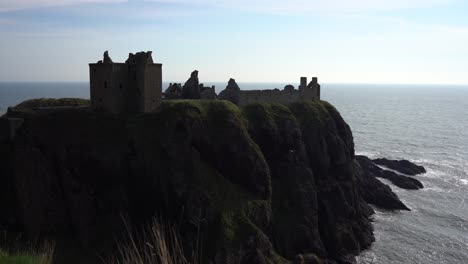 Majestuoso-Castillo-De-Dunnottar-En-Escocia-Encaramado-En-Un-Promontorio-Rocoso-Junto-Al-Océano,-El-Castillo-Está-Rodeado-Por-El-Rítmico-Rompimiento-De-Las-Olas-Mientras-Los-Elegantes-Pájaros-Vuelan-Por-El-Cielo