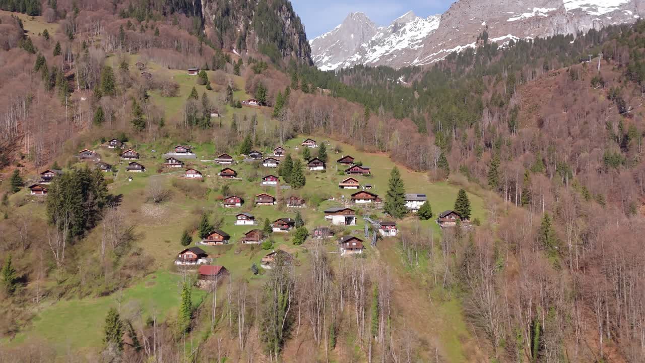 Alpebygda Schlattberg, Luchsingen, Sveits med tradisjonelle alpehytter spredt utover en bratt fjellside, omgitt av skog og snødekte fjell. Droneopptak fra luften.