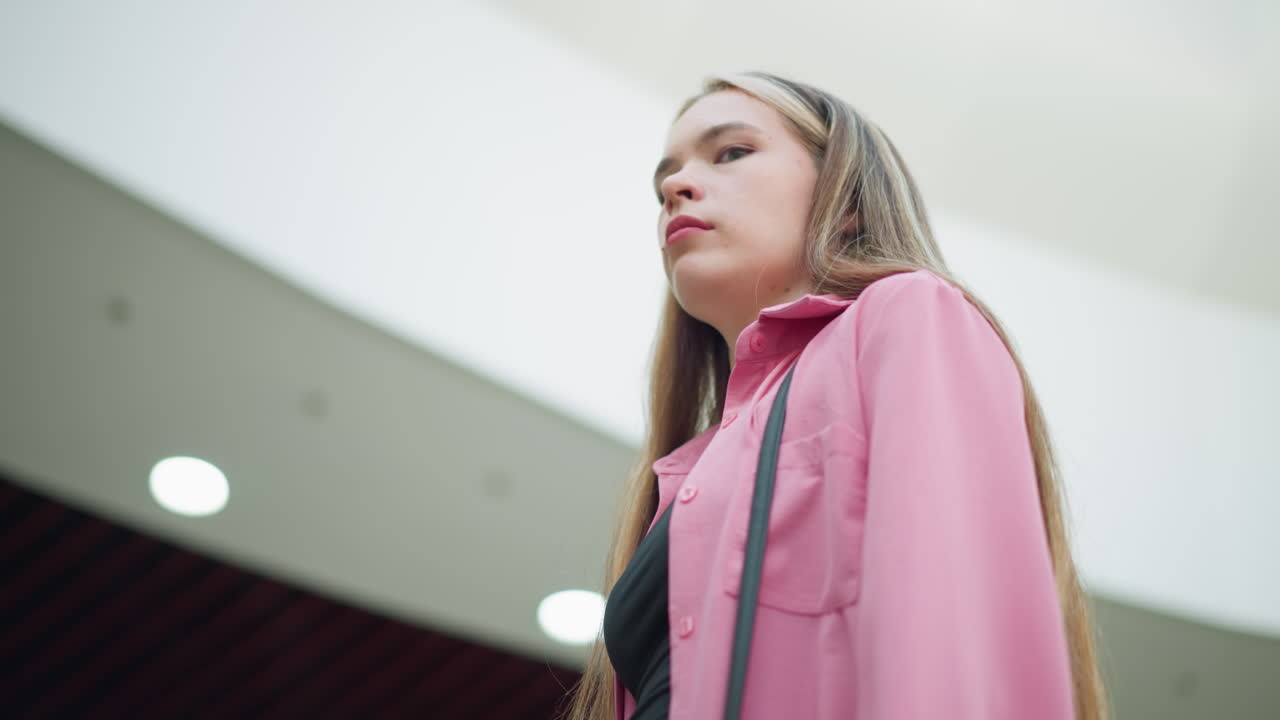 핑크색 드레스와 검은색 을 입은 젊은 여성은 실망한 표정으로 떠나기 전에 카메라로 사려 깊게 쳐다보고, 어깨에 크로스 보디 가방을 착용합니다.