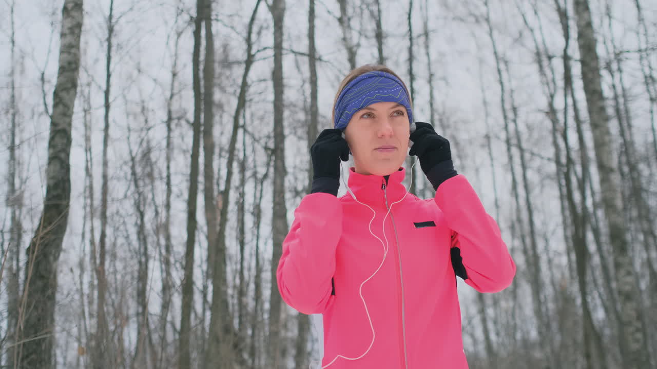 아침 겨울 조깅을 하기 전 소녀는 헤드폰을 귀에 꽂고 슬로우 모션으로 자연 공원을 달릴 준비를 하고 있습니다.