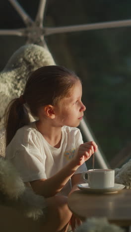 child stirs sweet tea sitting at table. peaceful girl looks at beautiful landscape behind transparent wall of glamping tent. happy vacation