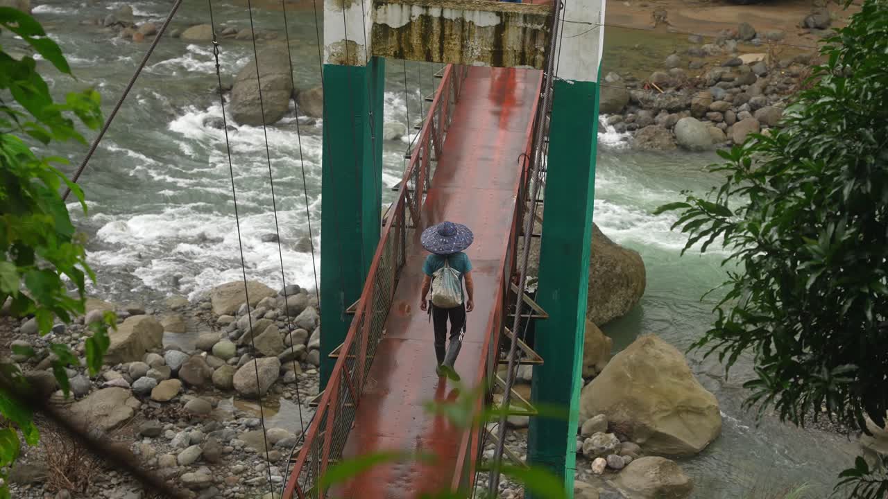 Captured from a riverside angle, two people cross a wet red footbridge over a rapid Philippine river, framed by lush greenery and misty forest air after rainfall