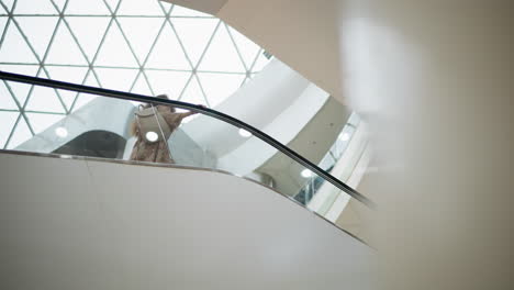 woman in vintage dress with handbag ascending on escalator, hand on rail, framed by modern glass and geometric ceiling design in bright mall environment