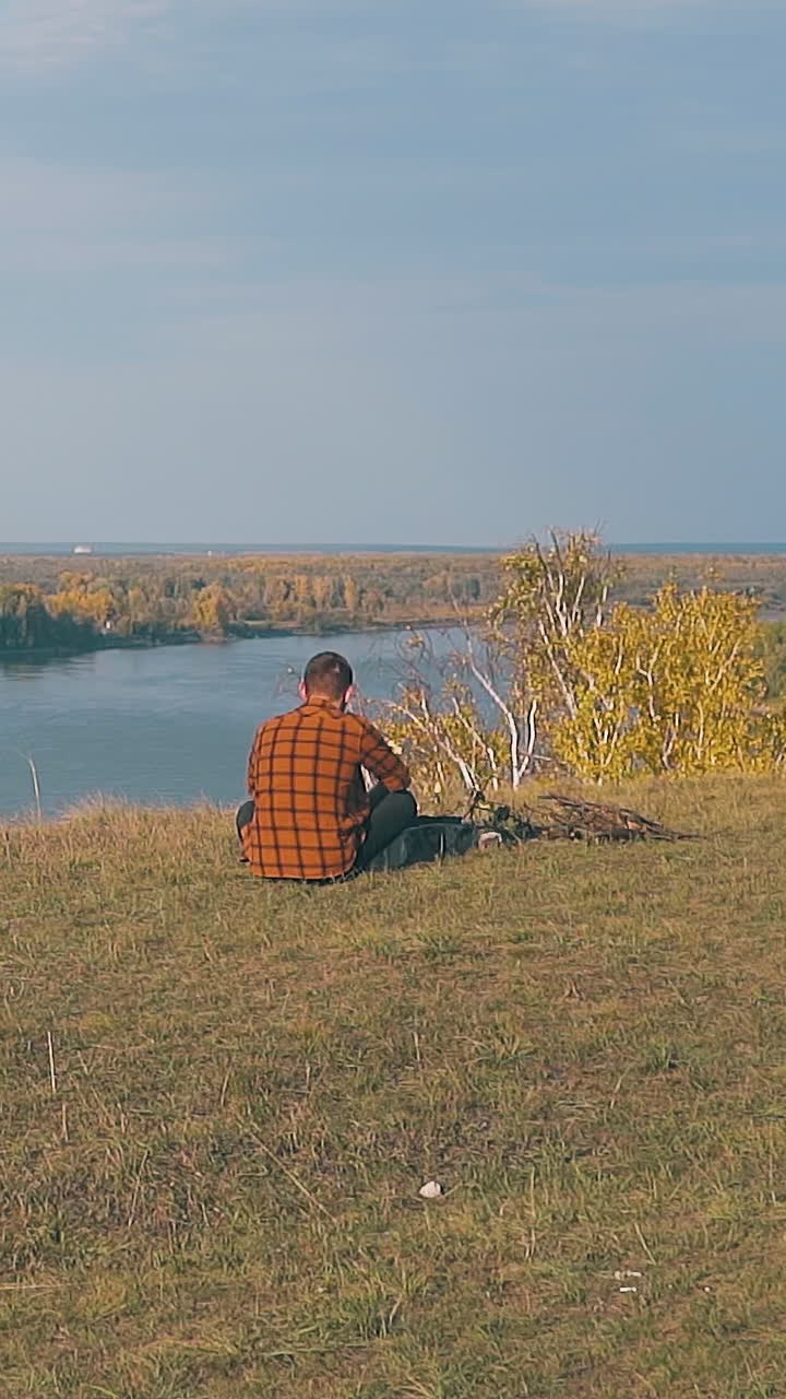 lonely young man sits at bonfire on meadow against wide calm river in early morning slow motion backside view