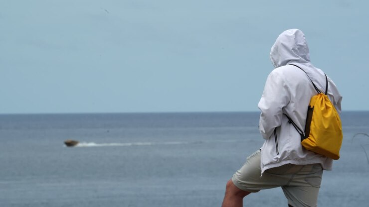 young male wearing hoodie and yellow rucksack back standing in front of the sea ocean while a boat cruise the seascape