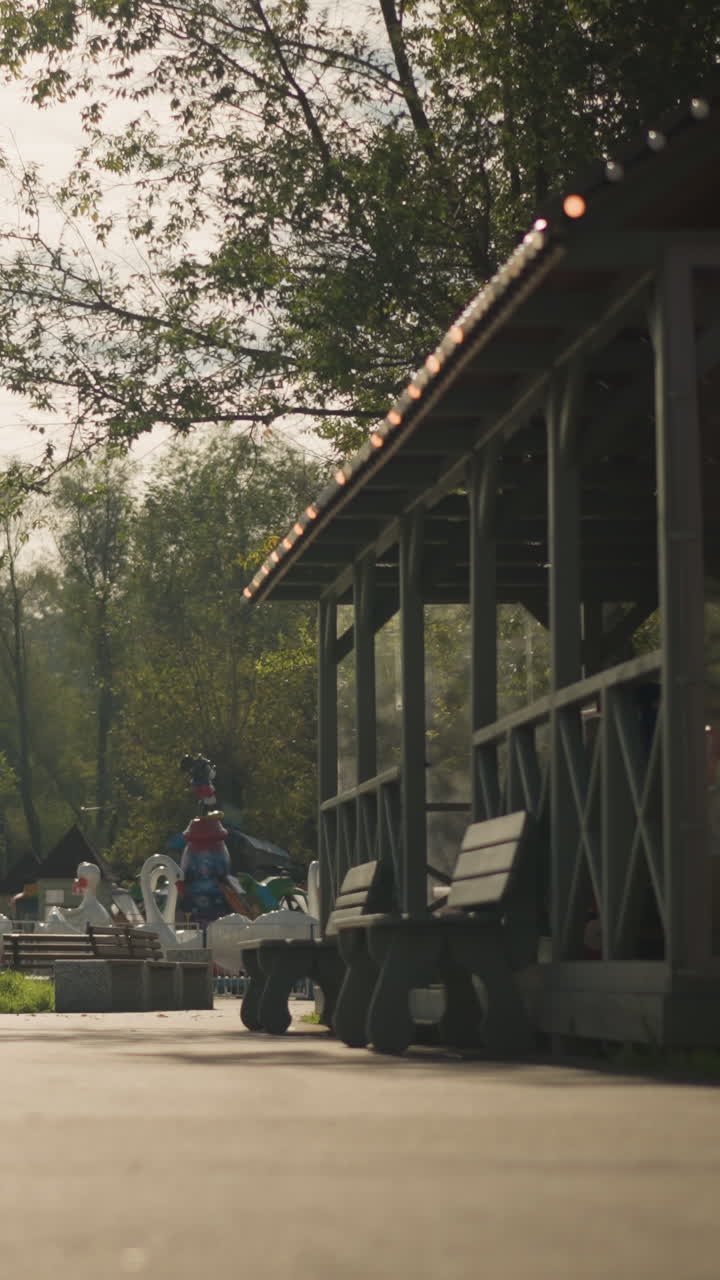 멀리 보이는 회전목마를 액자처럼 담은 나무 정자. 햇살이 비치는 통로, 빈 벤치, 잎이 무성한 덮개, 따뜻한 필름 톤, 느리게 움직이는 그림자, 은은한 보케, 놀이기구를 타는 멀리 있는 가족, 향수를 불러일으키는 여름 분위기, 소박한 느낌