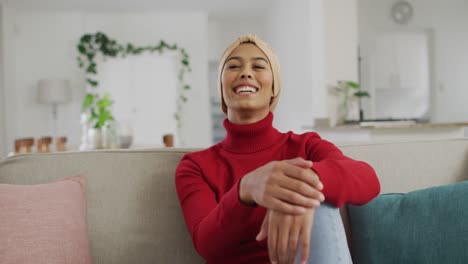 Video-of-happy-biracial-woman-in-hijab-sitting-on-sofa-and-looking-at-camera