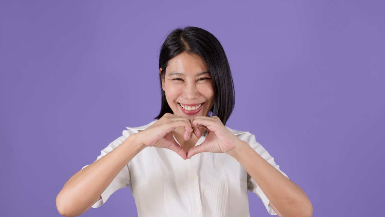 紫のスタジオ背景の前で、幸せそうなアジア人女性が手でハートの形を作っている。
