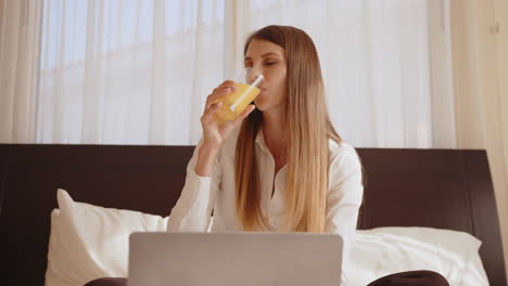 mujer trabajando desde la cama con computadora portátil y jugo de naranja
